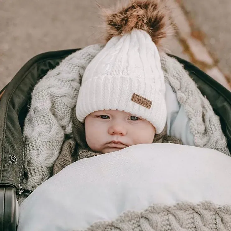 Baby med hvit lue med pompong i vognen i vognpose i grå kabelstrikk med hvitt fôr