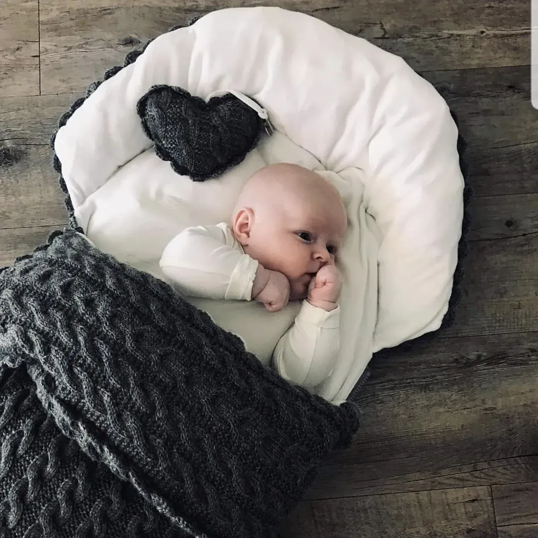 Baby ligger i et hvitt babyreir med teppe i mørkegrå kabelstrikk og et mørkegrått strikket hjerte