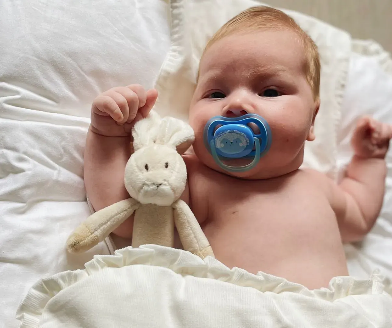 Baby med smokk og kosekanin ligger på en hvit dyne