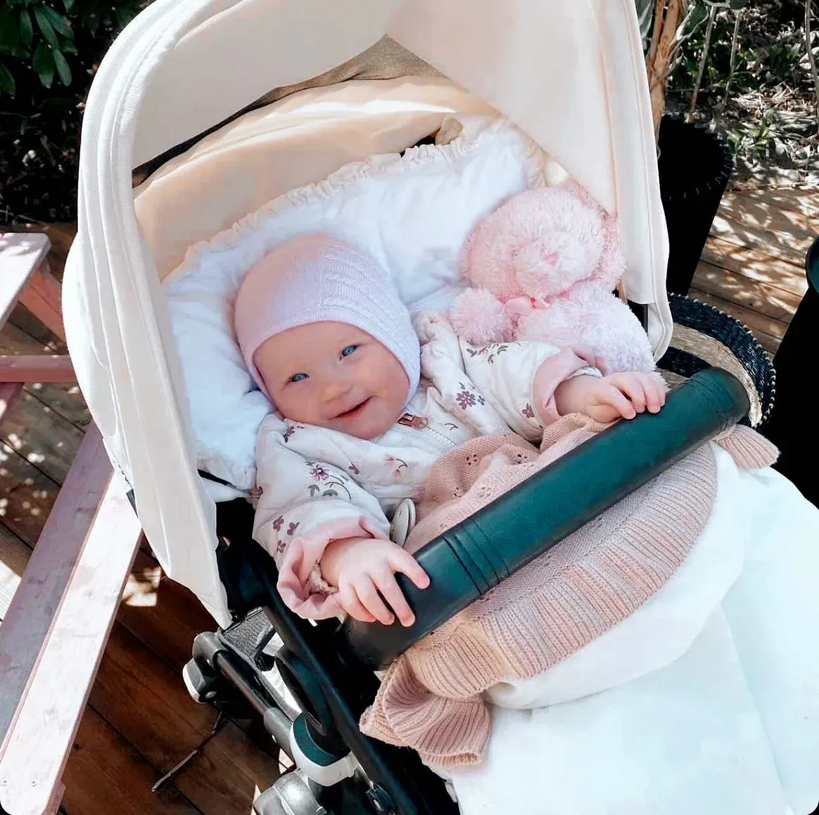 Baby i rosa lue smiler i en hvit barnevogn ute