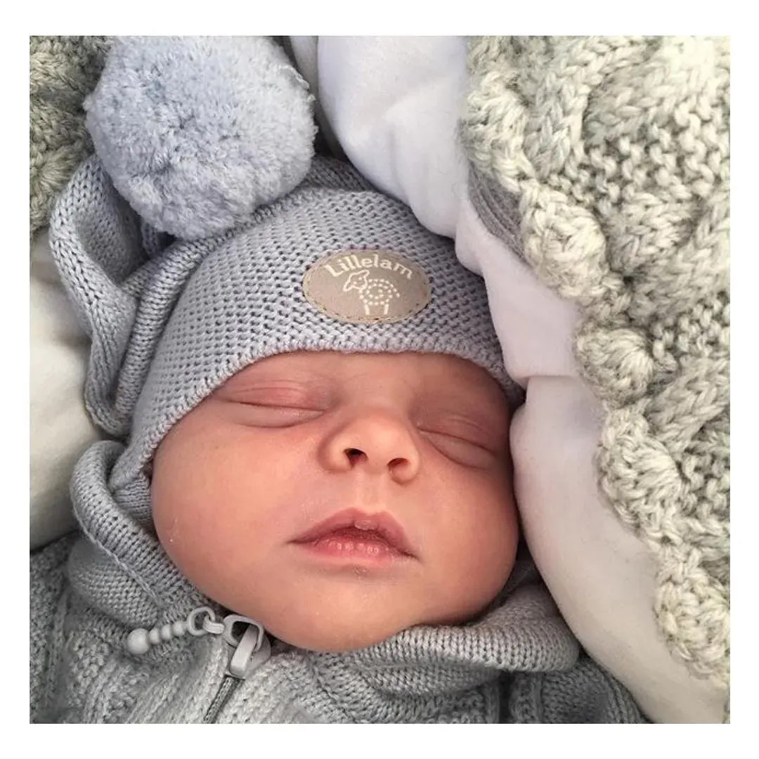 Baby i grå lue sover ved siden av kremhvit kabelstrikk