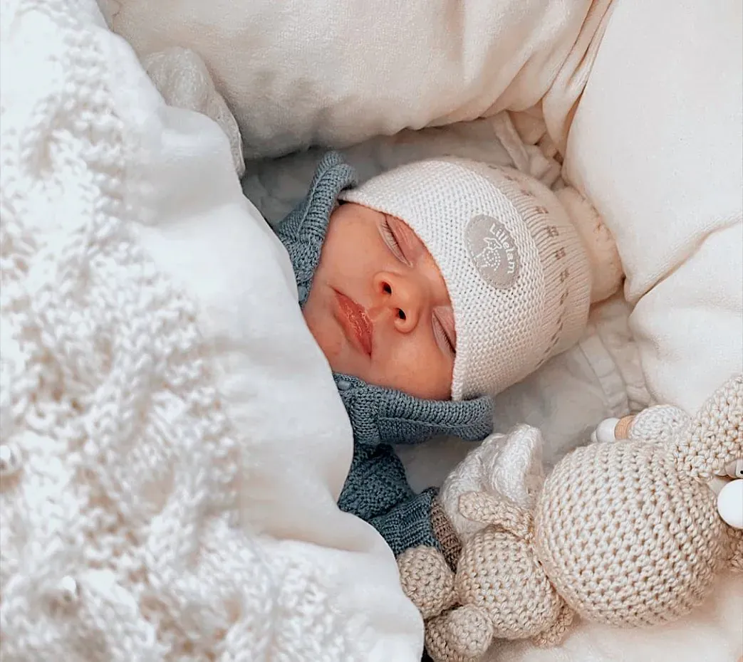 Baby sover i hvit lue under kremhvit strikk med et heklet kosedyr