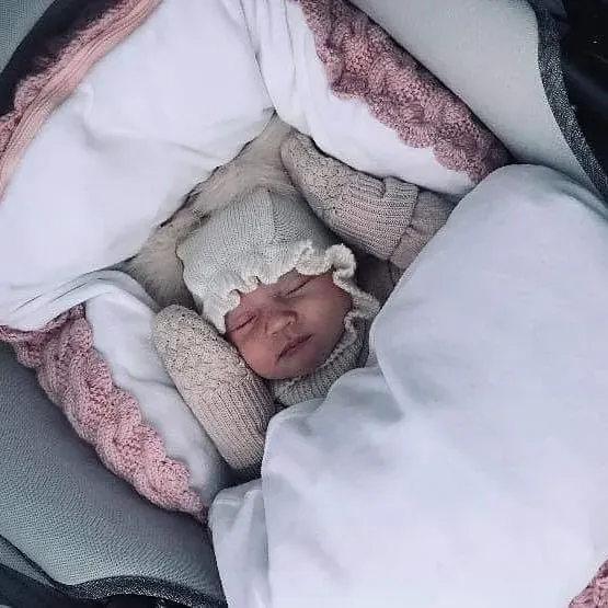 Baby i strikkelue sover i vognen i en hvit pose med kant i rosa kabelstrikk