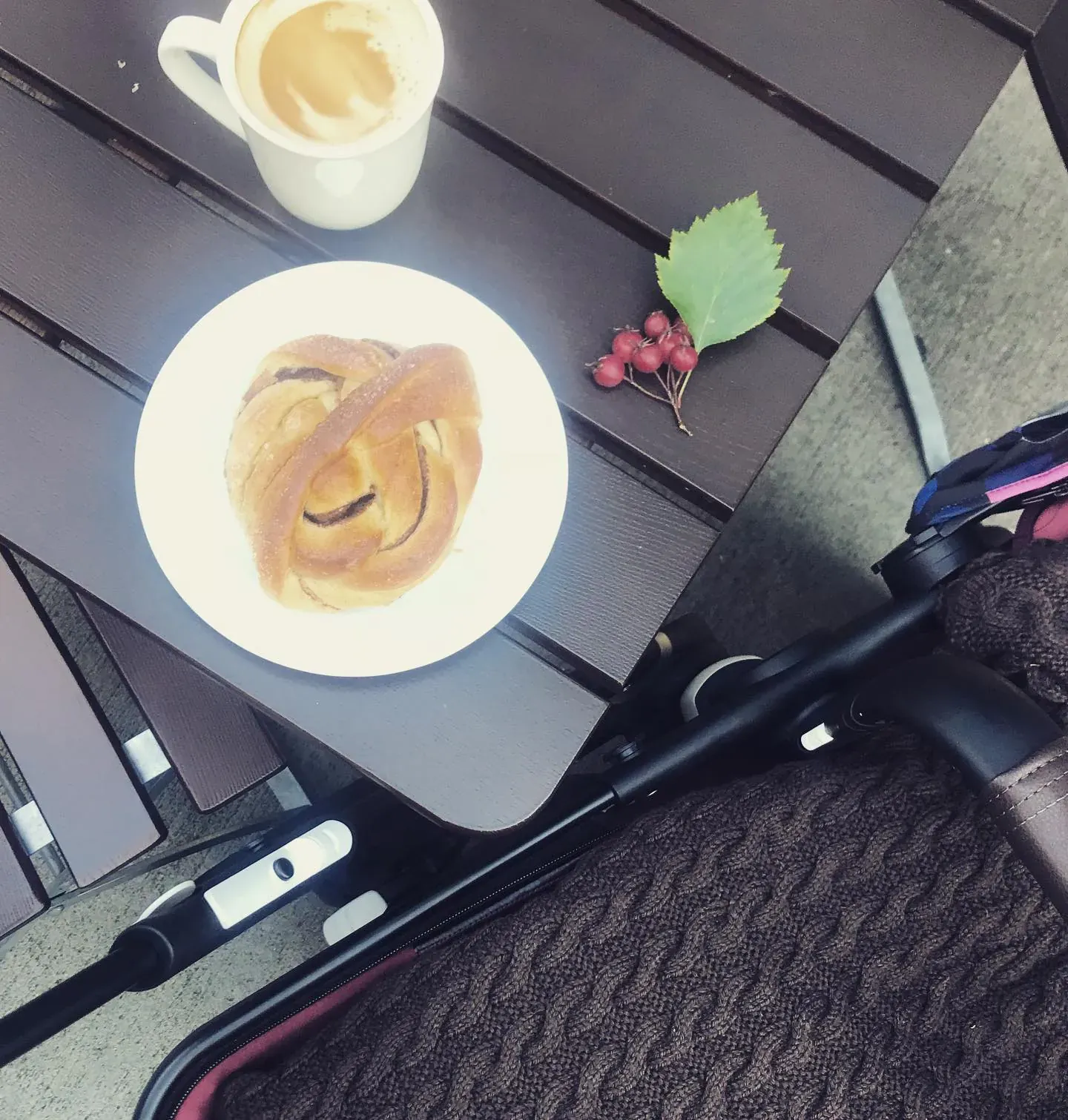 Kaffe og bolle på et bord ved siden av en barnevogn med vognpose i brun kabelstrikk