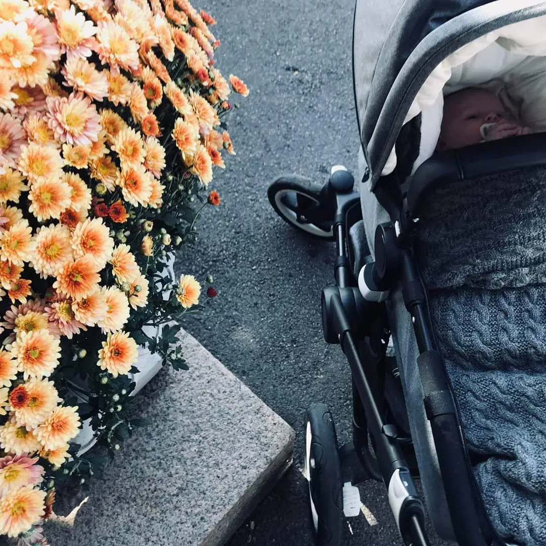 Barnevogn med vognpose i grå kabelstrikk ved siden av oransje blomster