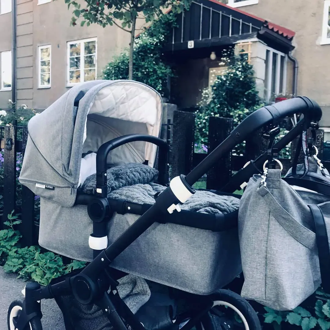 Grå barnevogn med vognpose i grå kabelstrikk parkert utenfor et hus