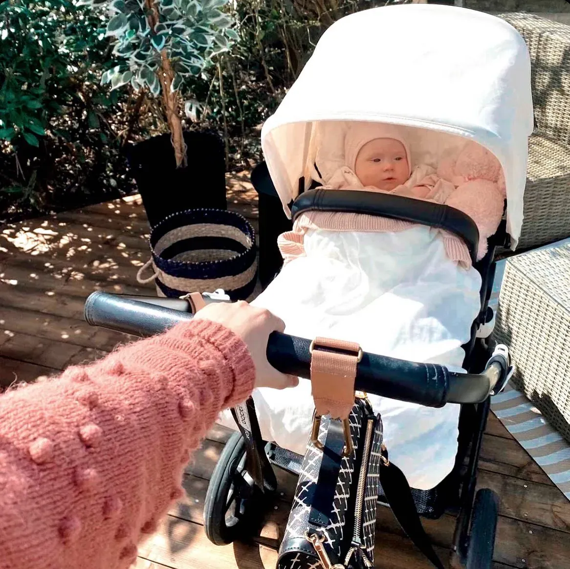 Hånd i rosa strikkegenser holder en hvit vogn med baby i hvit pose på en terrasse