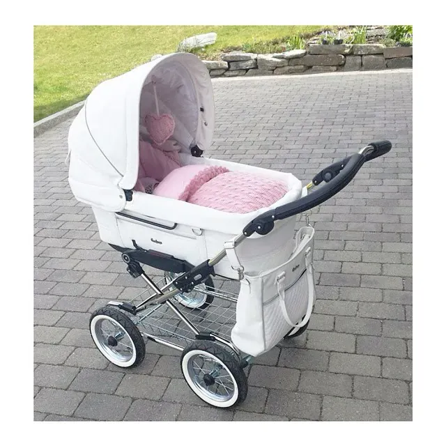 Hvit barnevogn ute med en vognpose i rosa kabelstrikk og et rosa vognhjerte