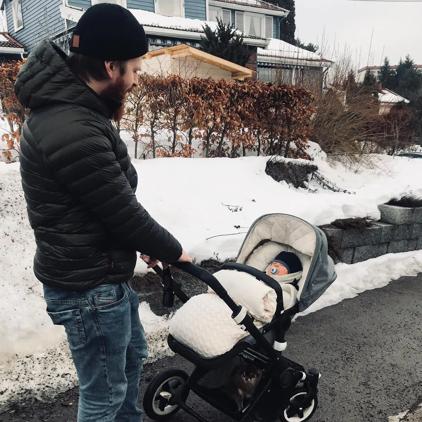 Mann triller barnevogn med vognpose i kremhvit kabelstrikk ute i snøen