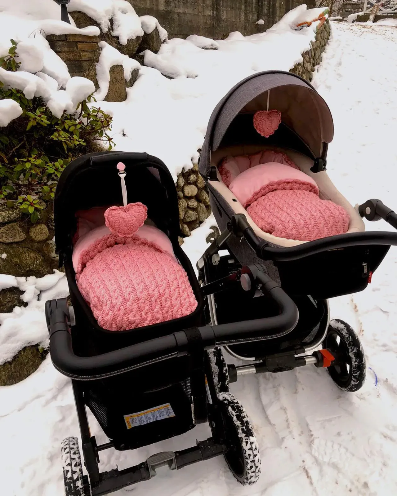 To svarte vogner med vognposer i rosa kabelstrikk og strikkede vognhjerter ute i snøen