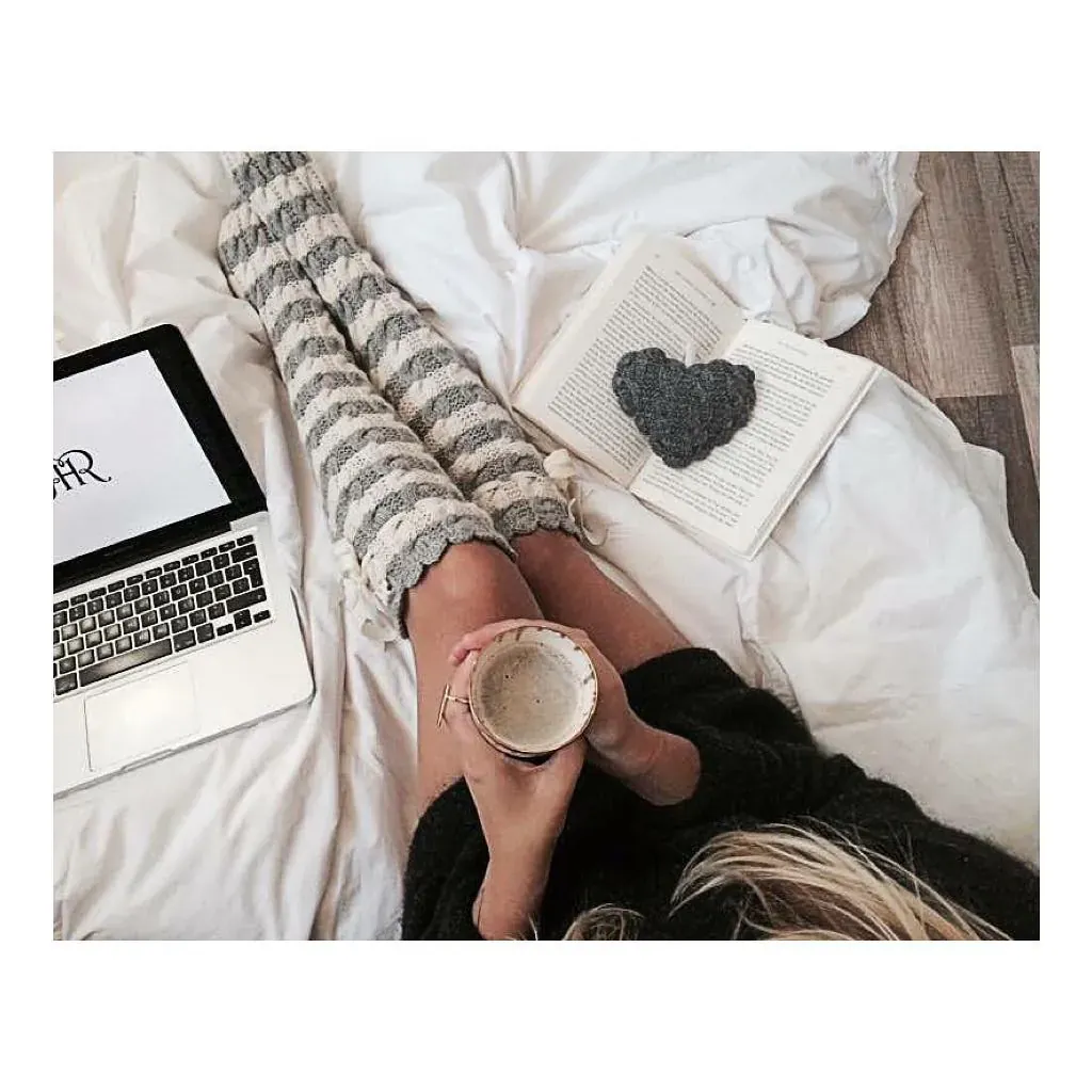 Kvinne i sengen med kaffekopp, laptop og en bok med et grått strikket hjerte
