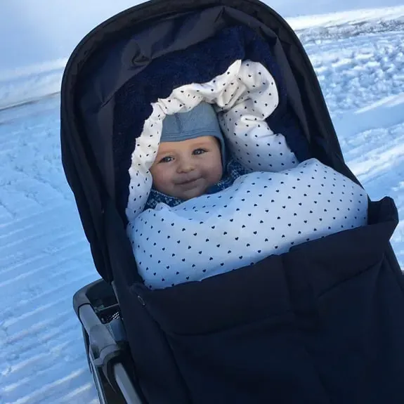 Baby smiler i en svart vogn med hvit pose med prikker, ute i snøen