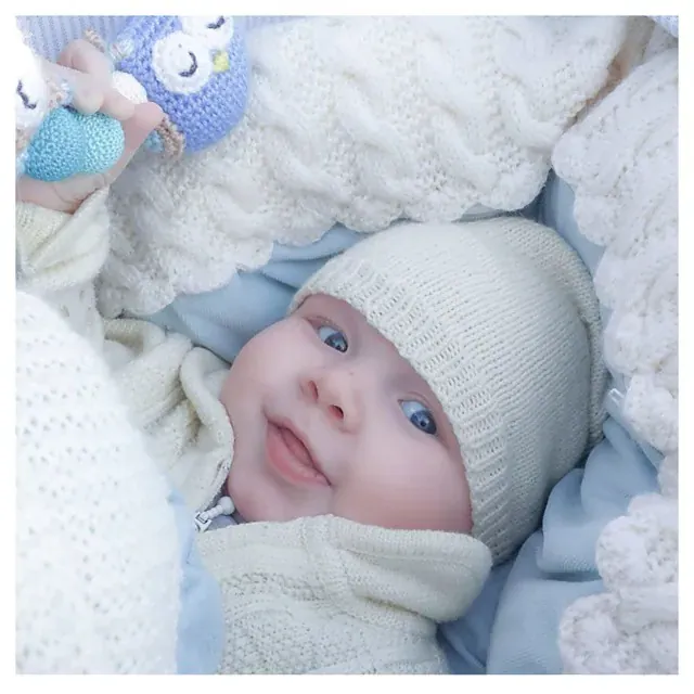 Baby med hvit strikkelue i en vognpose i hvit kabelstrikk