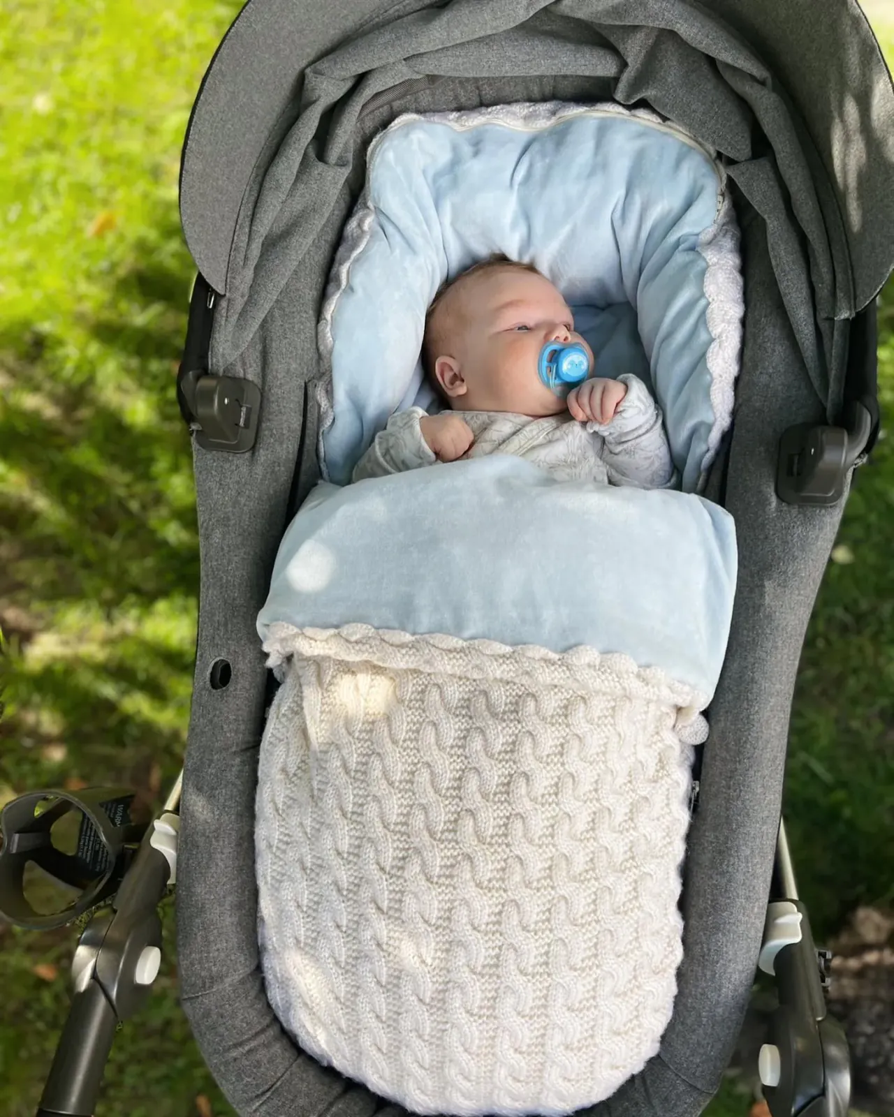 Baby med smokk i vognpose i hvit kabelstrikk med lyseblått fôr i en grå vogn