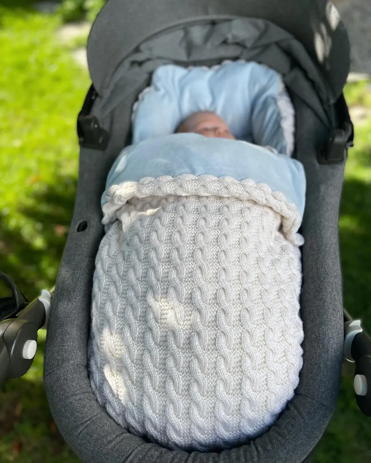 Vognpose i hvit kabelstrikk med lyseblått fôr i en grå vogn på gresset