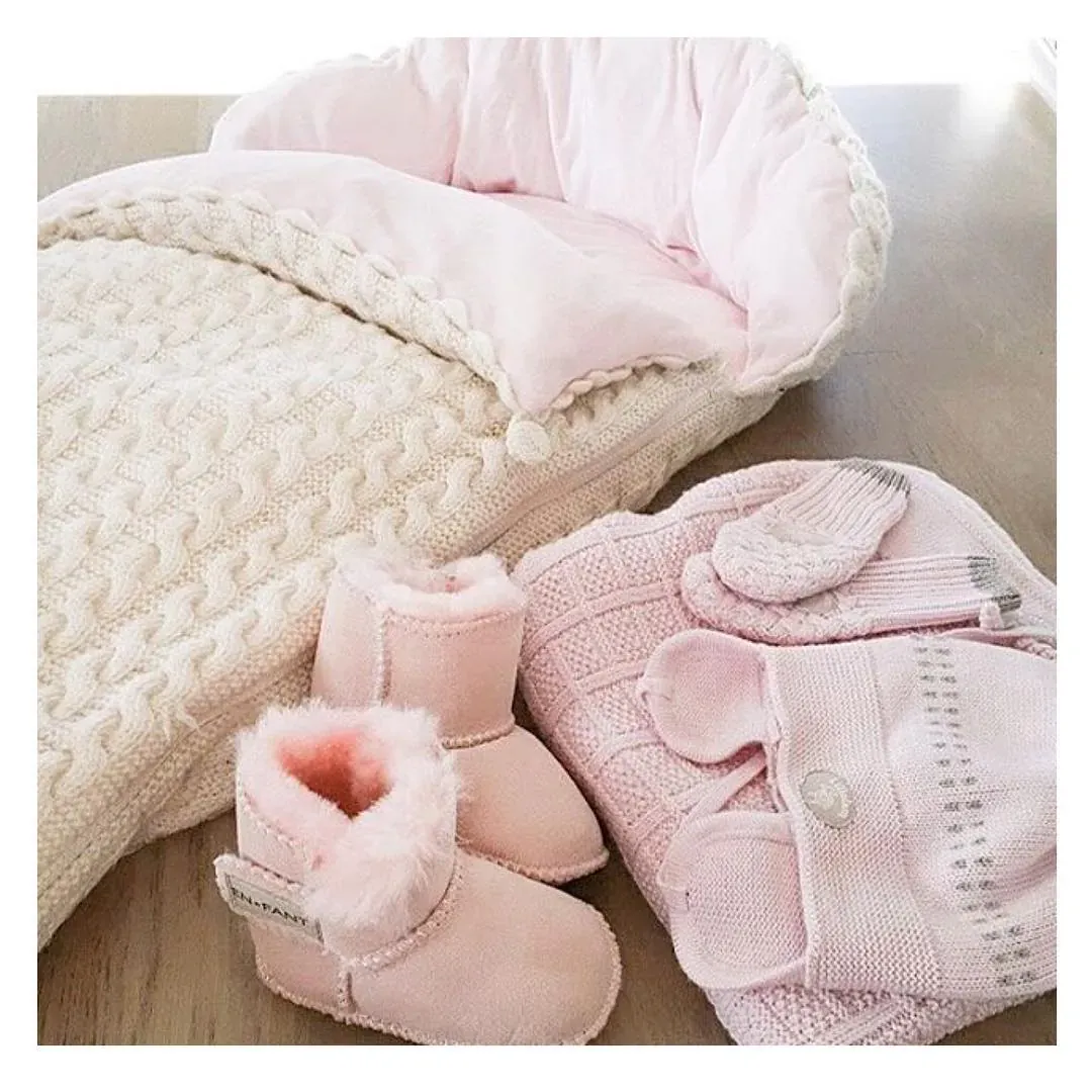 Vognpose i kremhvit kabelstrikk med rosa fôr ved siden av rosa babytøfler og strikkejakke