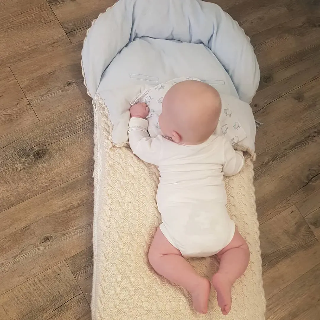Baby ligger på magen på en vognpose i kremhvit kabelstrikk med lyseblått fôr, sett ovenfra