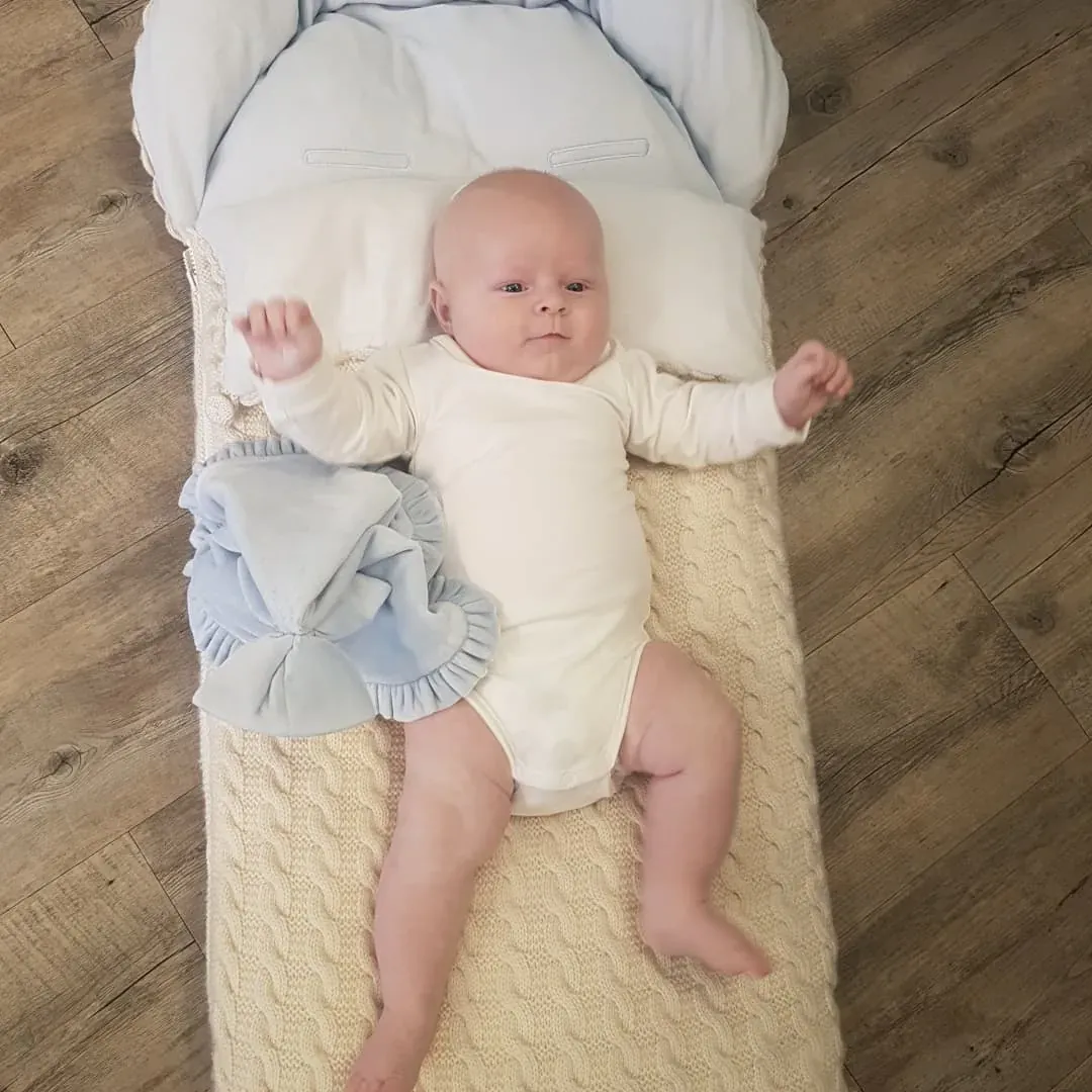 Baby ligger på ryggen på en vognpose i kremhvit kabelstrikk med lyseblått fôr