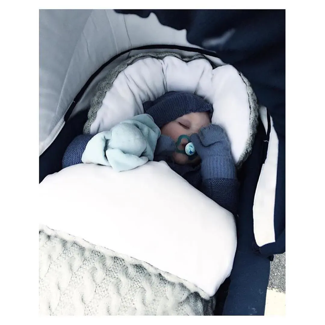 Baby med smokk og lue i vognen, i vognpose i lysegrå kabelstrikk med hvitt fôr