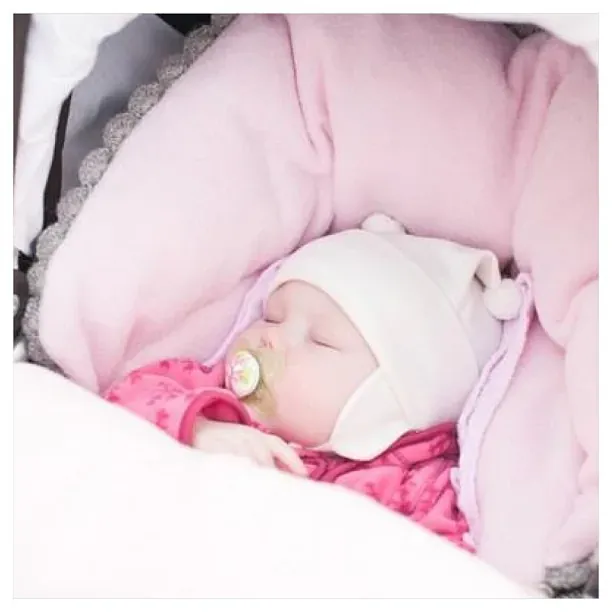 Baby med hvit lue og smokk sover i en vognpose med rosa fôr og kant i grå strikk