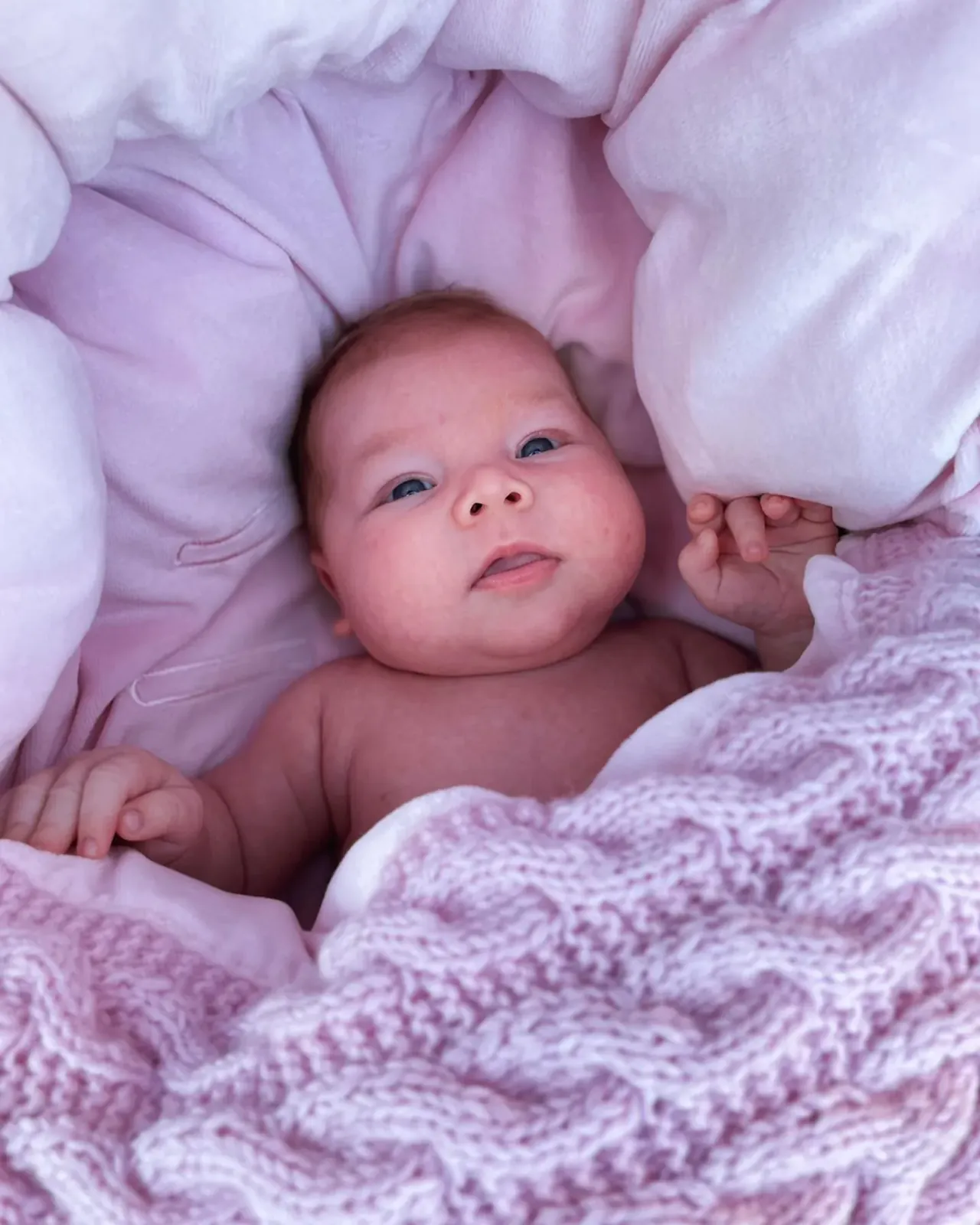 Baby ser opp fra vognpose i rosa kabelstrikk