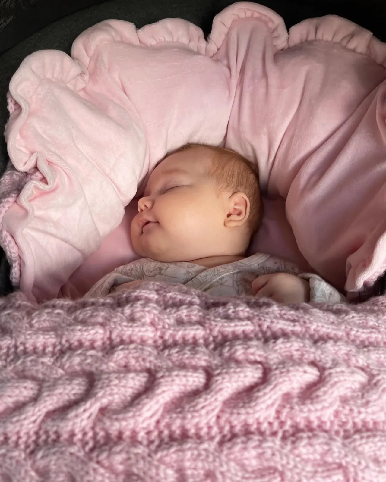 Baby sover i vognpose i rosa kabelstrikk med rosa fôr og store rysjer