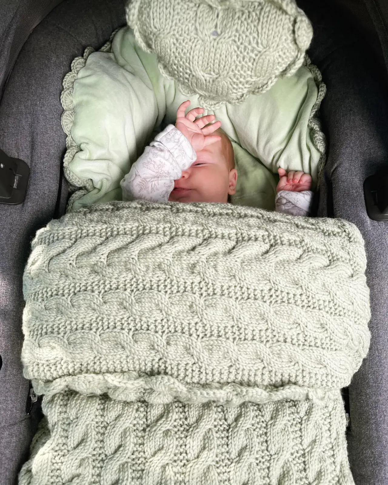 Baby sover i vognen i en vognpose i salvie kabelstrikk med grønt fôr
