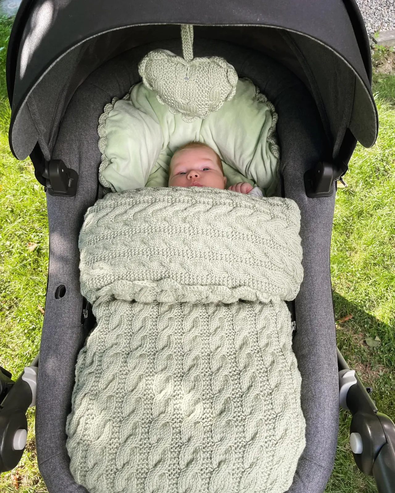 Baby i vognpose i salvie kabelstrikk med lysegrønt fôr og et strikket vognhjerte over