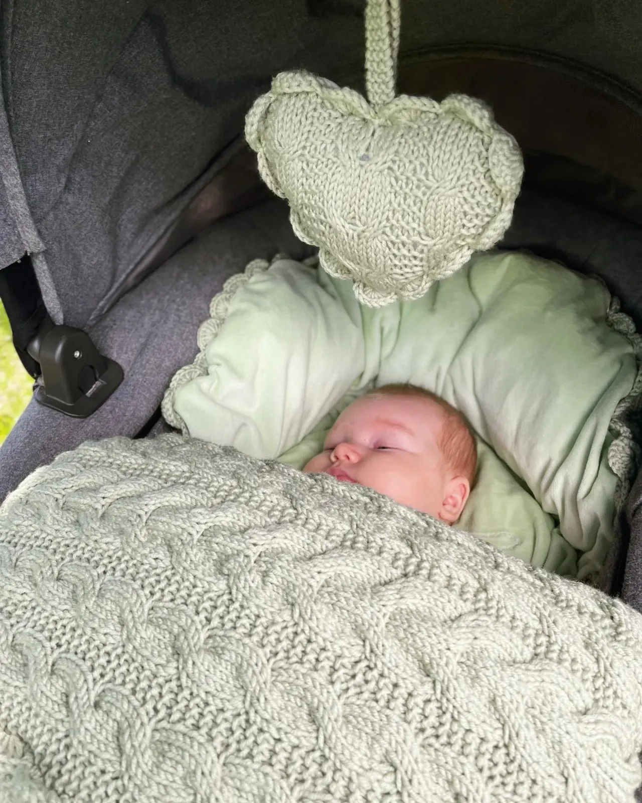 Baby sover i vognpose i salvie kabelstrikk med lysegrønt fôr under et strikket vognhjerte